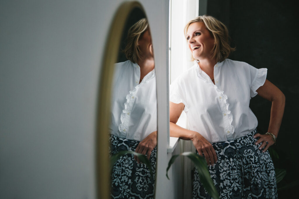 Woman in a white blouse and colorful pants looking out the window.  Photography by Lindsay Hite