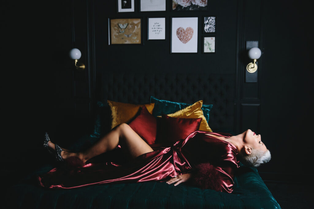 Woman in satin maroon robe laying on back on a teal jewel-toned bed.  Photography by Lindsay Hite