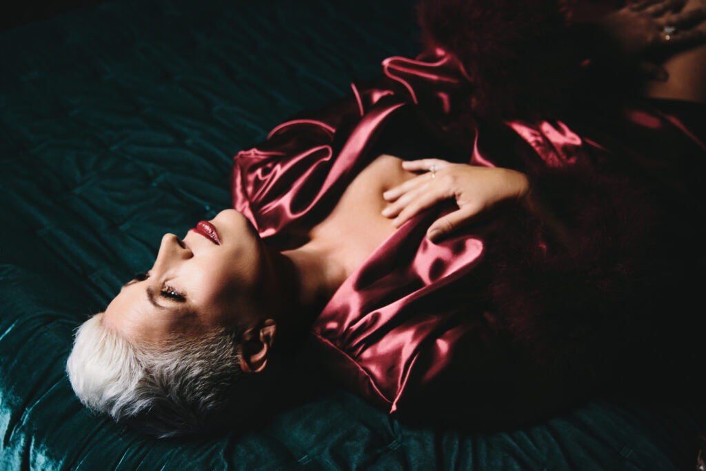 Woman in rose satin rob laying on her back on a teal jewel-toned bed with her hand on her chest. Photography by Lindsay Hite