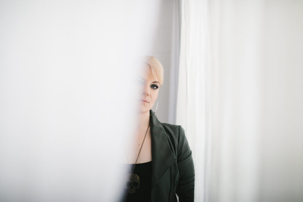 Woman in black leather jacket halfway behind a white curtain.  Photography by Lindsay Hite