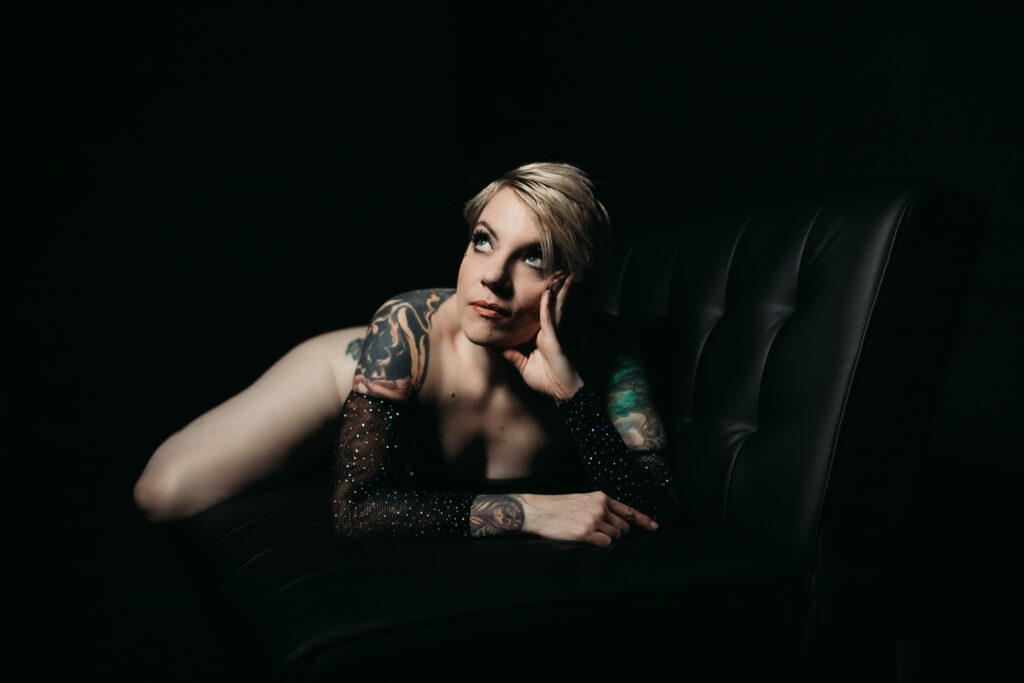 Woman in black rhinestone body suit laying on a black couch with a black backdrop. Photography by Lindsay Hite. 