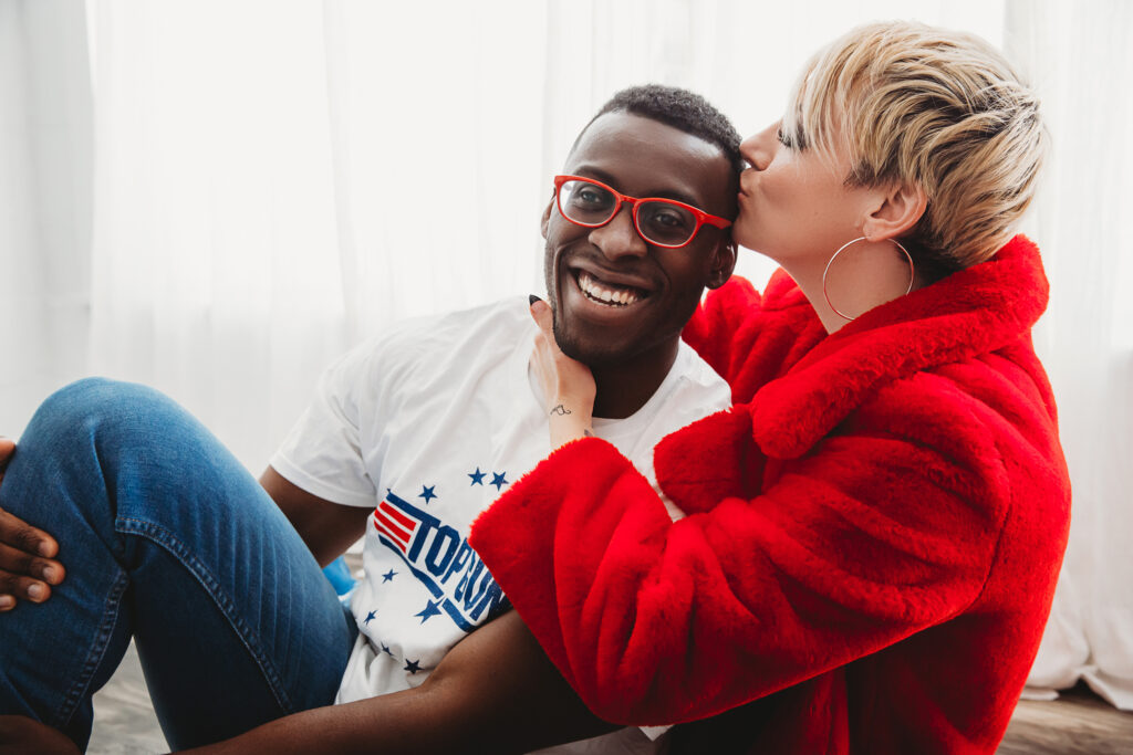 Woman in bright red fleece jacket kissing her partner on the head while embracing laughter and love. Photography by Lindsay Hite.