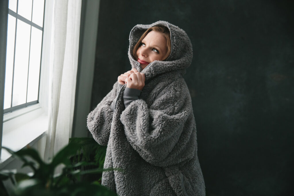 Woman in fleece hoodie blanket looking out the window with a smile on her face.  Photography by Lindsay Hite