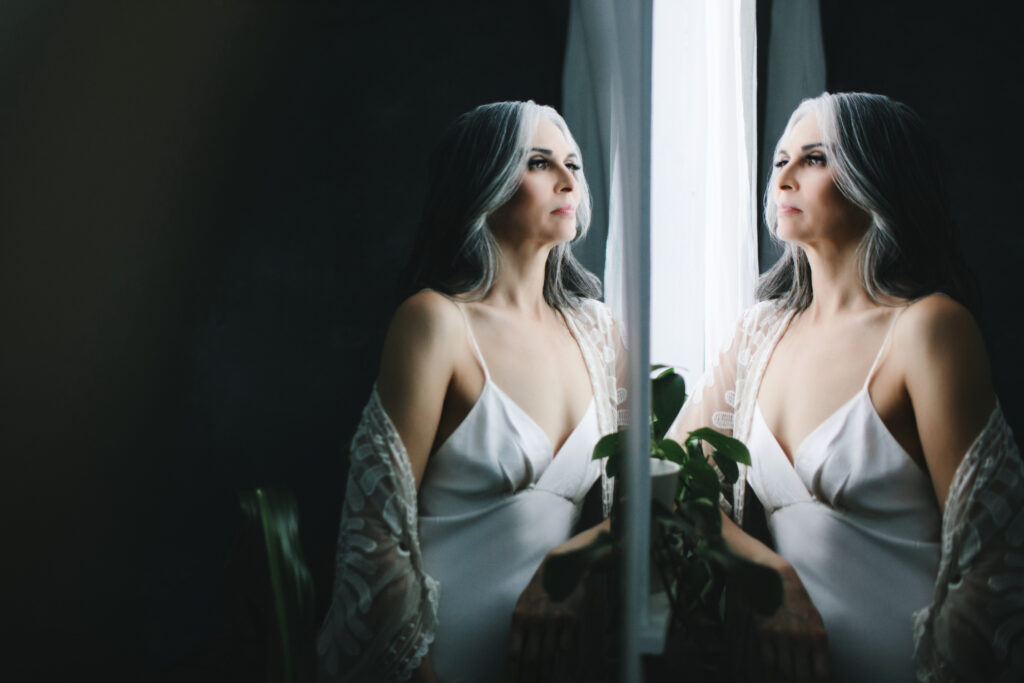 Woman in white slip dress and shawl looking out the window.  Photograph by Lindsay Hite. 