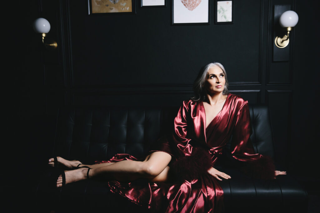 Woman in long dark blush silk robe sitting on a black sofa looking out in the distance.  Photography by Lindsay Hite