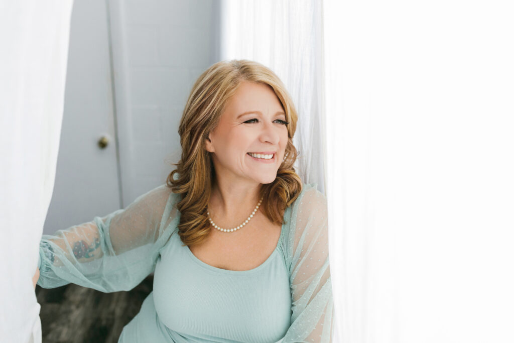 Woman in pale teal shirt and pearls looking out the window with joy on her face knowing her worth. Photography by Lindsay HIte