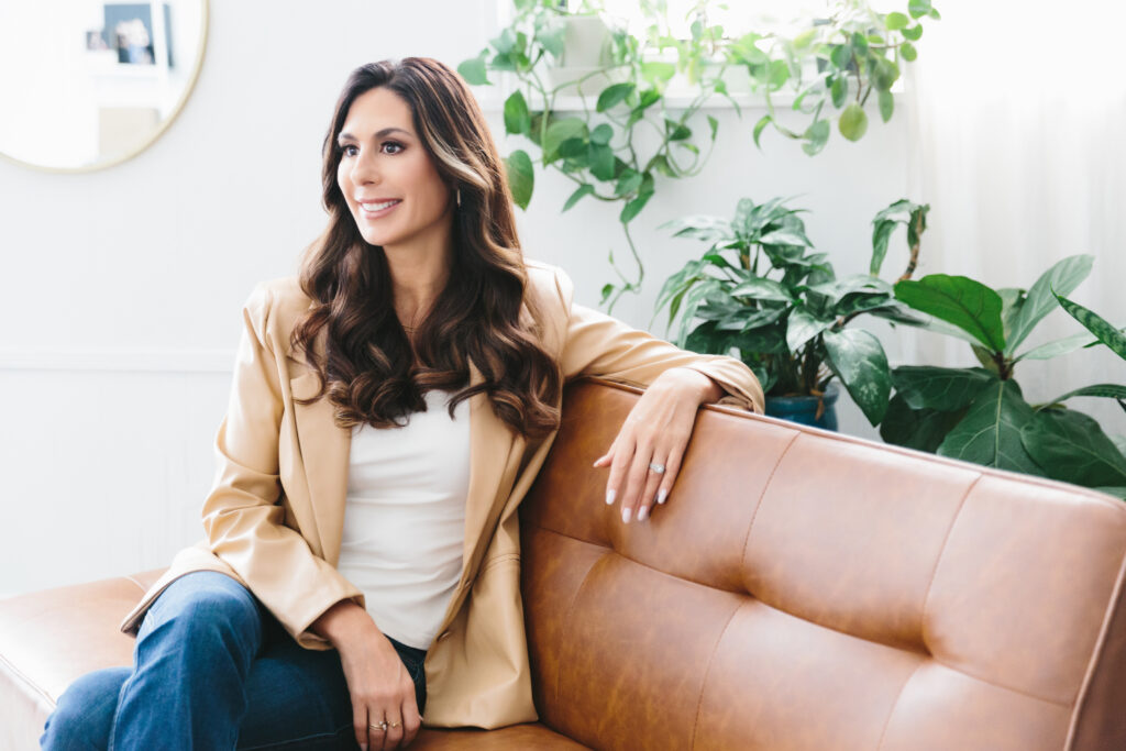 Woman in a white shirt, jeans, and a tan blazer sitting on a tan sofa with plants in the background. redefining health and success for women over forty. Photography by Lindsay Hite.