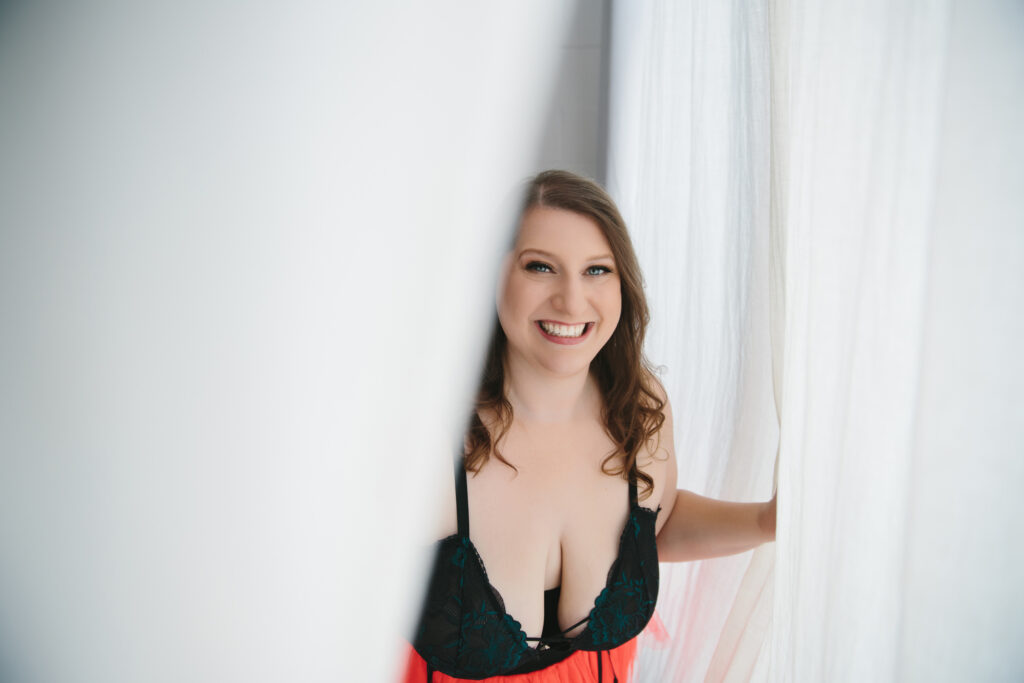 Woman in black top and red skirt looking through curtains with a smile on her face. Photography by Lindsay Hite