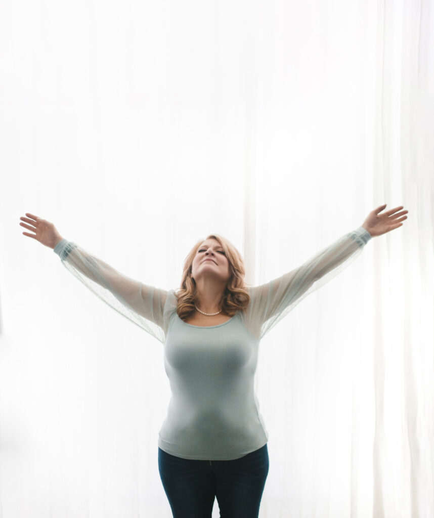 Woman in pale teal shirt looking up at the sky with her arms spread out wide in a star pose against a white curtain background.  Photography by Lindsay Hite
