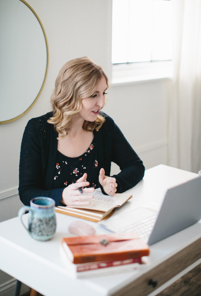 Woman wearing a navy flowered top with navy cardigan, sitting at a desk with a journal in her hand in a Zoom call. Photography by Lindsay Hite