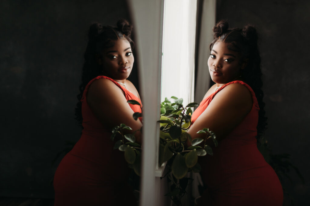 A woman in an orange dress leaning against a windowsill looking at the camera with her reflection showing in the mirror.  Photography by Lindsay Hite