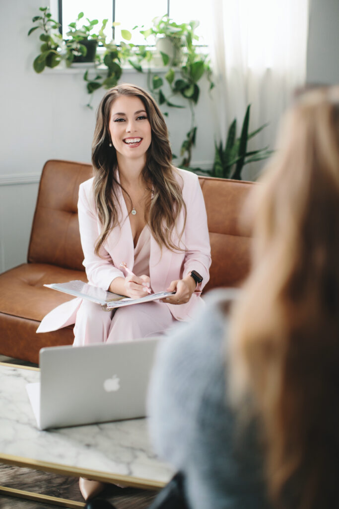 Woman in a pink suit taking notes in her notebook with her laptop open while speaking with a client. Photography by Lindsay Hite
