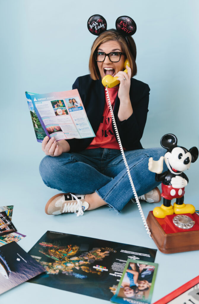 Woman wearing jeans, a red shirt and black jacket with a Mickey Mouse hat, holding a yellow phone to hear ear and a Disney brochure in her other hand with Disney items surrounding her. Photography by Lindsay Hite