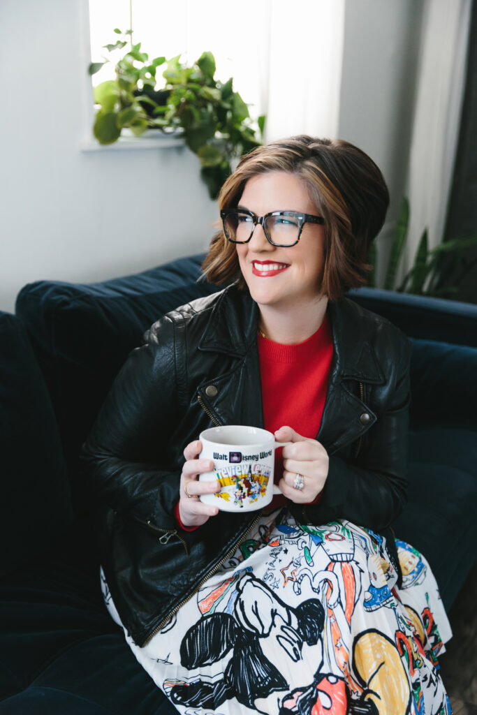 Woman wearing a Disney-inspired skit, red shirt, and black leather jacket sitting on a navy sofa and holding a Disney coffee mug while looking off in the distance. Photography by Lindsay Hite.