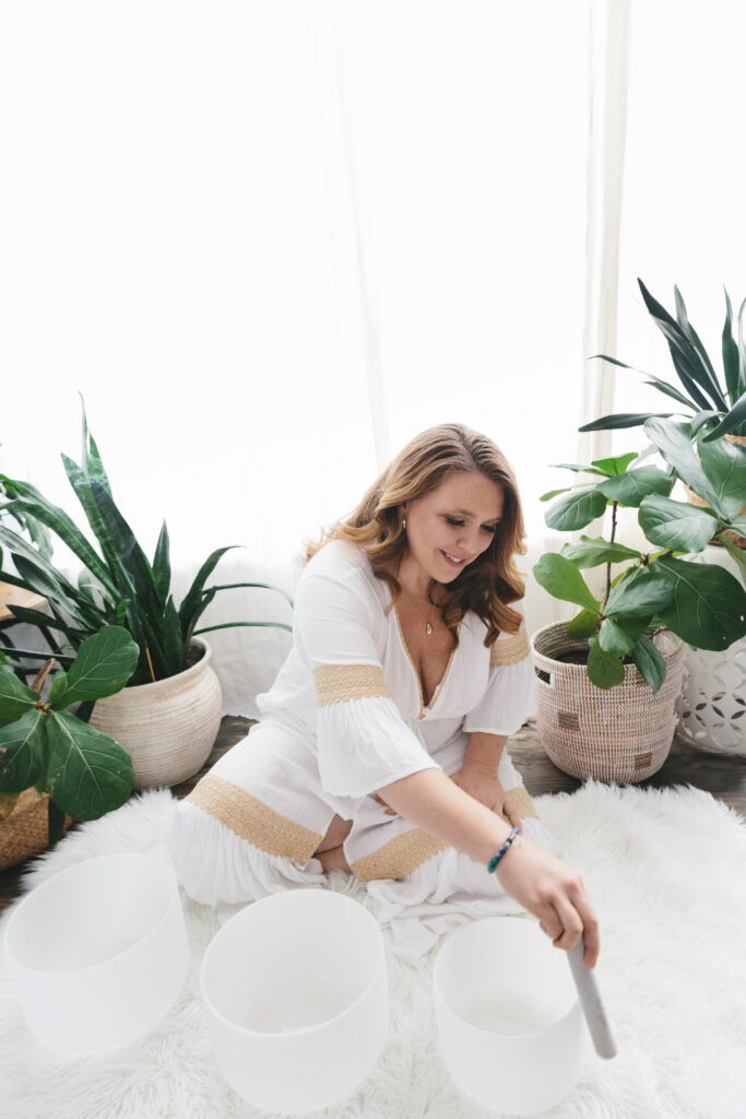 Woman in linen white dress sitting on a white fur rug playing crystal sound bowls. Photography by Lindsay Hite