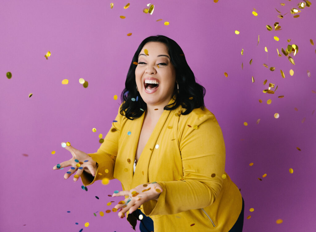 Woman in yellow jacket against a pinkish purple background throwing confetti in the air. Photography by Lindsay Hite