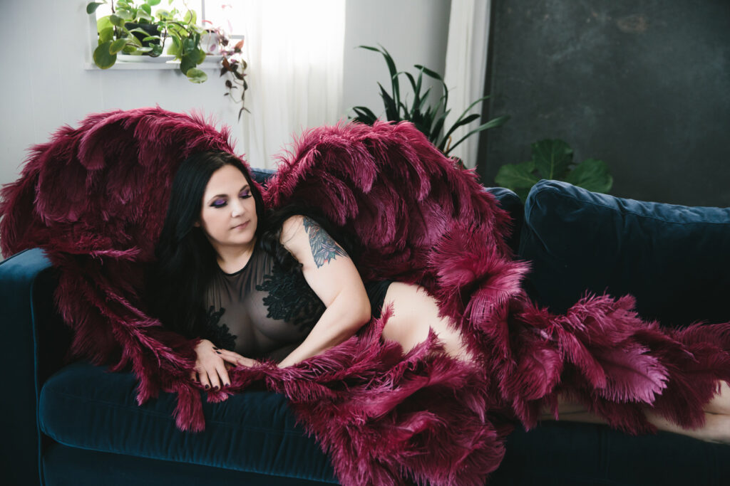 Woman in black lingerie wearing burgundy angel wings sitting on a navy blue sofa.